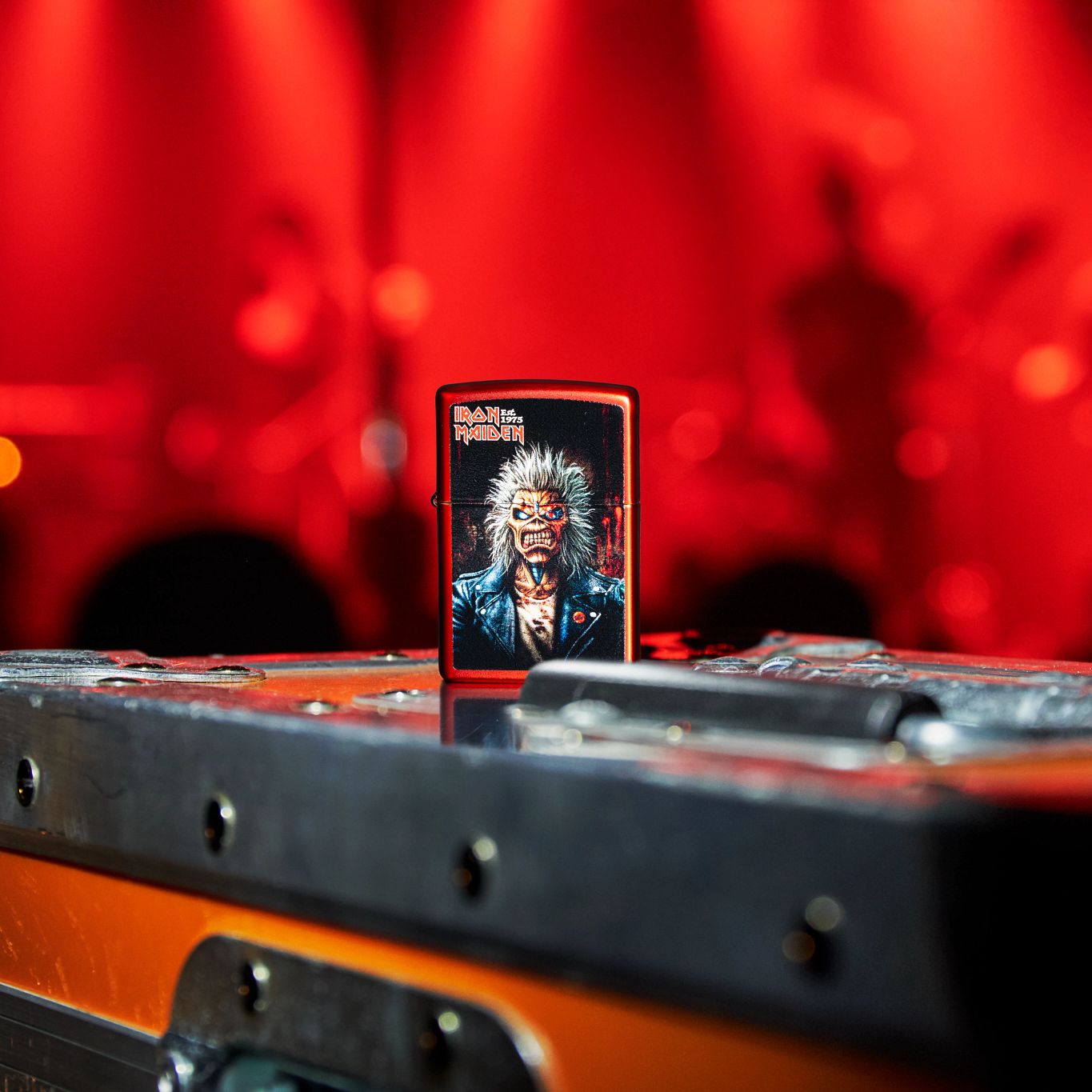 Lifestyle image of Zippo Iron Maiden 50th Metallic Red Windproof Lighter standing on a chest in front of a blurred concert scene.