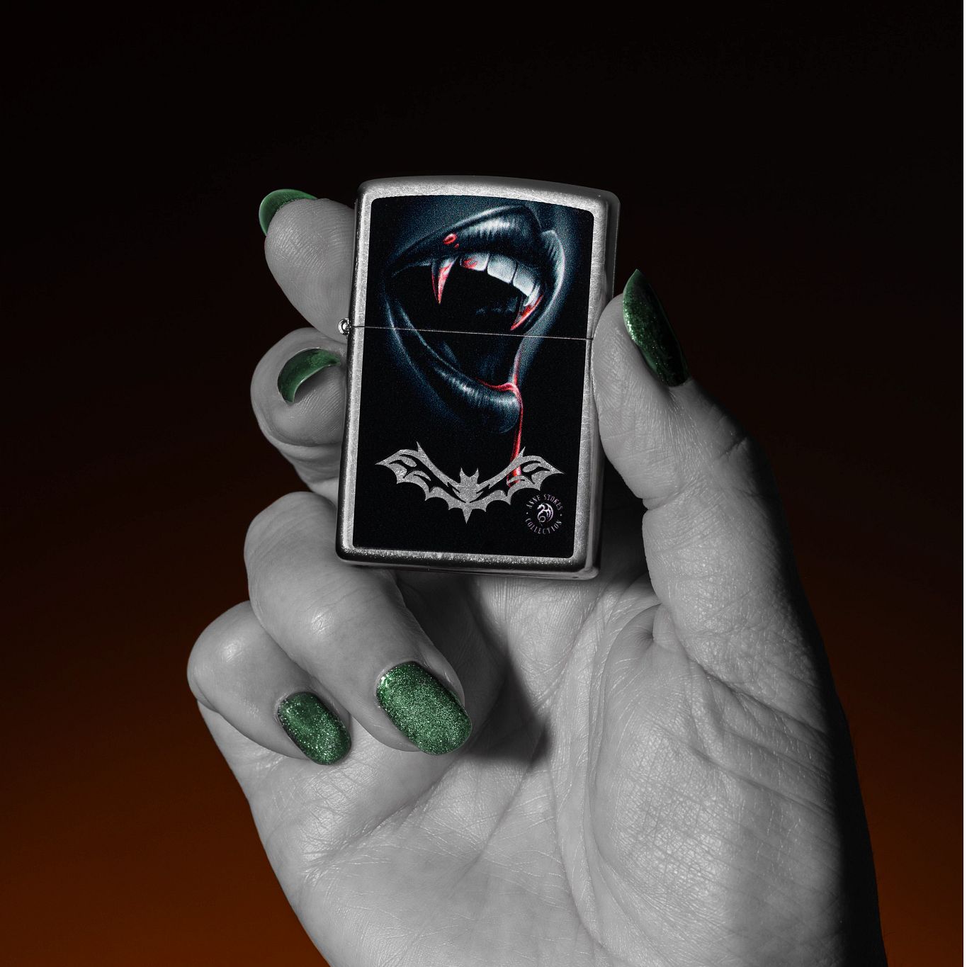 Lifestyle image of Zippo Anne Stokes Bite Design Street Chrome Windproof Lighter being held in a hand with a black and orange ombre background. 

