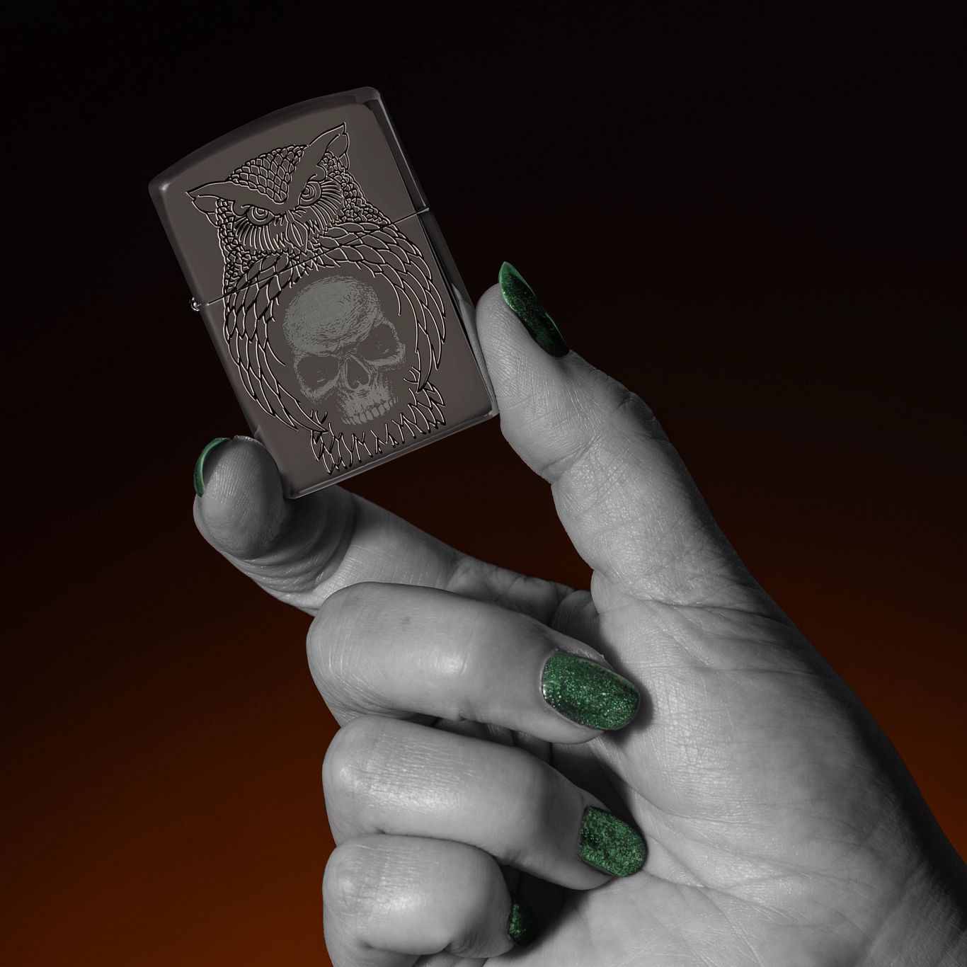 Lifestyle image of Zippo Night Owl Design Armor Black Ice Windproof Lighter being held in a hand with a black and orange ombre background.