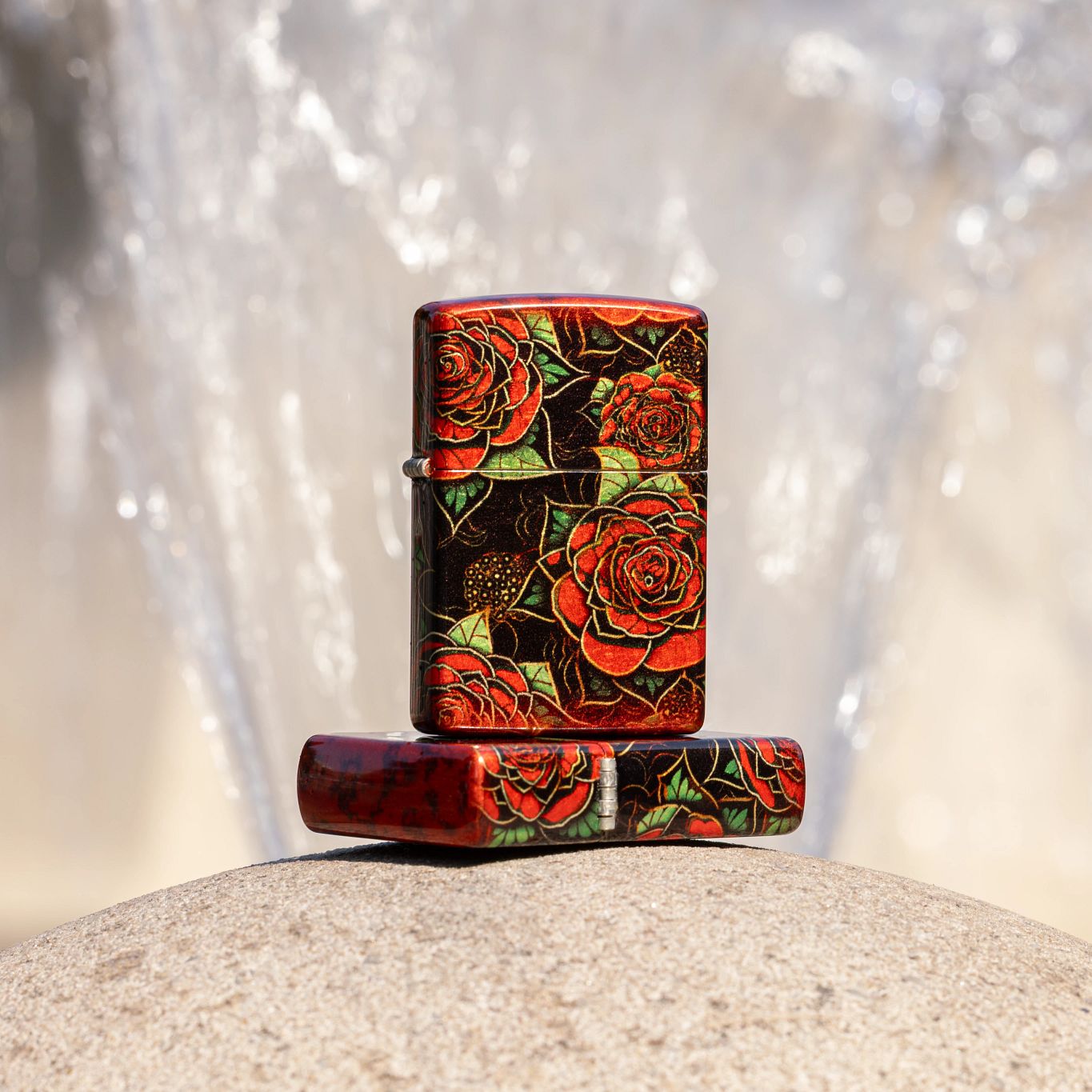 Lifestyle image of two Zippo Blooming Roses Design 540 Tumbled Brass Windproof Lighters; one lying flat on a rounded light tan concrete surface showing the top of the lid and hinge side of the lighter while the other lighter is standing on top of the lighter that is lying flat showing the front of the lighter with a blurred fountain with water running down as the background. 

