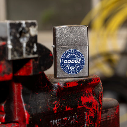 Lifestyle image of Zippo Dodge Service Sign Street Chrome Windproof Lighter standing on an old mechanics pro vise with a blurred garage in the background. 

