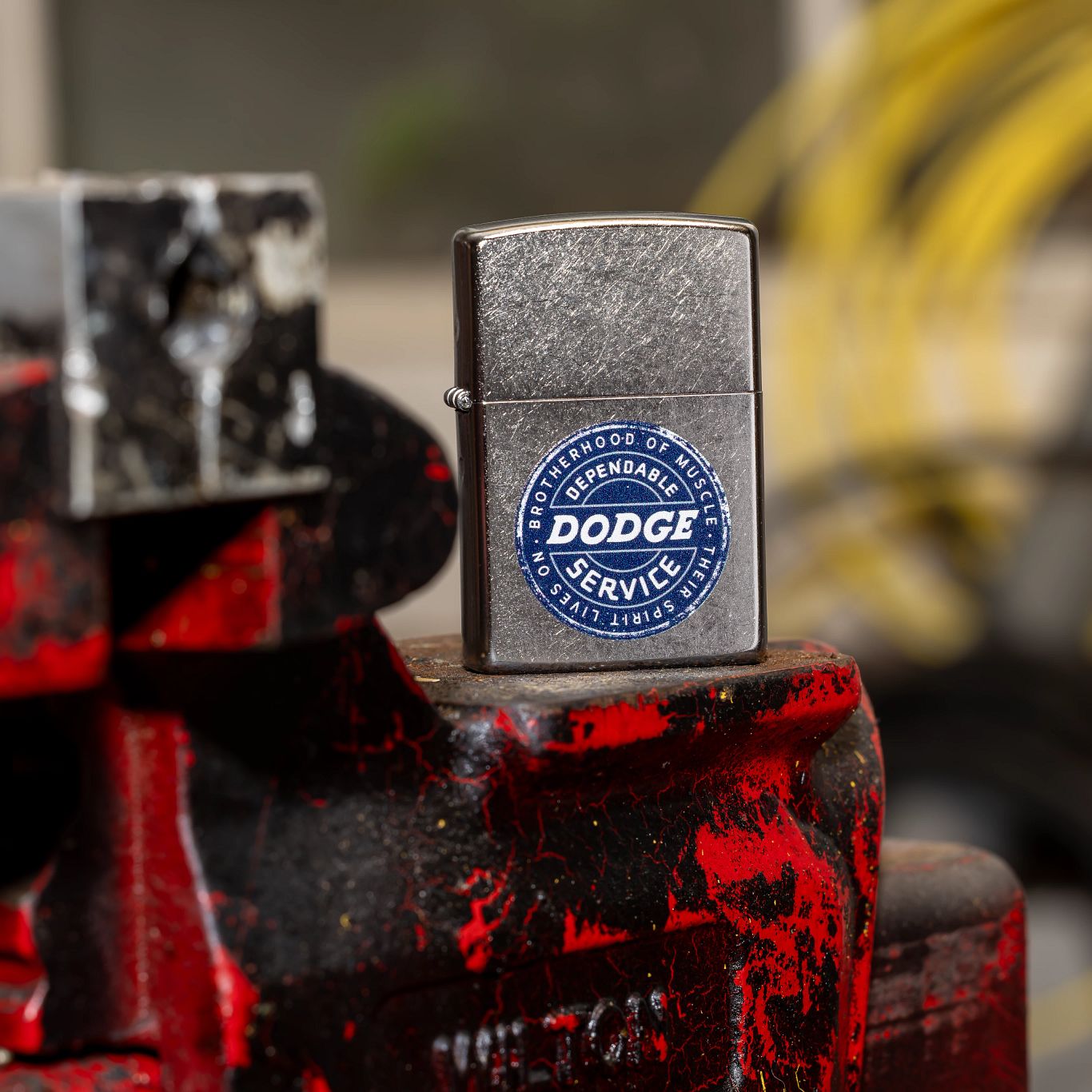 Lifestyle image of Zippo Dodge Service Sign Street Chrome Windproof Lighter standing on an old mechanics pro vise with a blurred garage in the background. 

